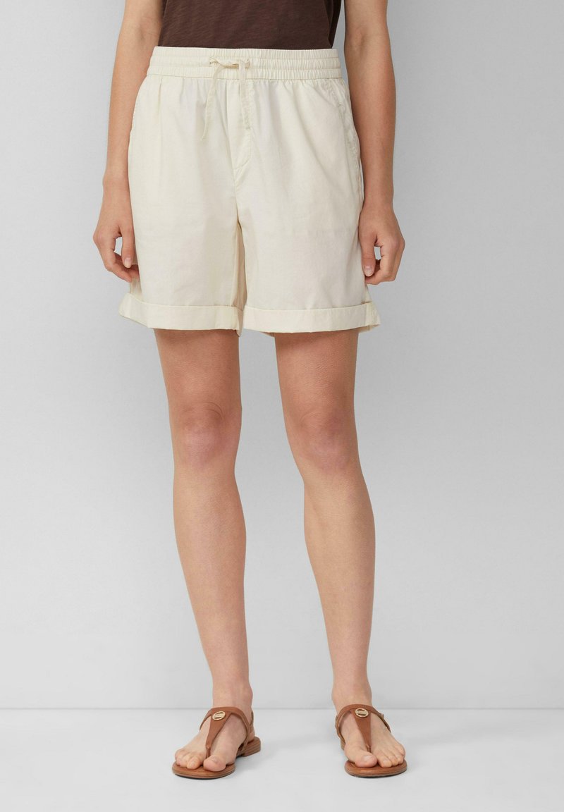 Pantalones cortos de algodón beige claro con cintura elástica, cordón ajustable y dobladillos enrollados. Llevados con sandalias de tiras marrones.