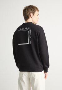 Fekete pulóver, kerek nyakkivágással és hosszú ujjakkal. Hátul "Calvin Klein" felirat és egy fehér téglalap grafika látható.