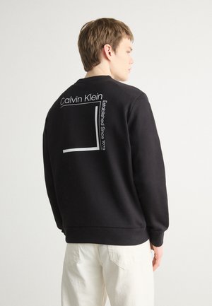 Juodas megztinis su apykakle ir ilgomis rankovėmis. Nugarinėje pusėje yra užrašas "Calvin Klein" ir stačiakampio formos grafika balta spalva.