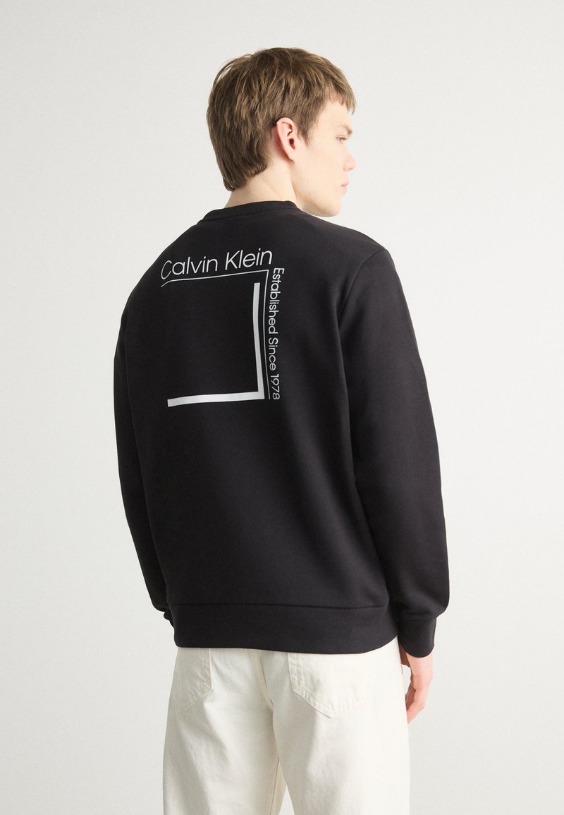 Fekete pulóver, kerek nyakkivágással és hosszú ujjakkal. Hátul "Calvin Klein" felirat és egy fehér téglalap grafika látható.