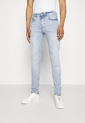 Beine in hellblauen, eng anliegenden Jeans und weißen Turnschuhen, stehend vor einem schlichten weißen Hintergrund.