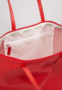 Raudona nešiojamoji rankinė su tekstūruotu paviršiumi, dvigubomis rankenomis ir balta vidine dalimi. Yra užtrauktuko kišenė ir spausdintas 'LACOSTE' logotipas krašte.