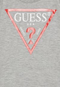 Szürke pamut póló, amelyen egy nagy piros háromszög grafika látható a "GUESS" logóval, "U.S.A." felirattal és fehér "WASHED JEANS" szöveggel.