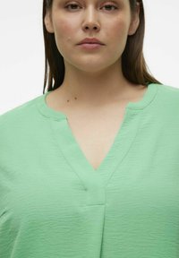 Blouse vert menthe avec un décolleté en V, texture douce et détail de couture subtil. Présente une coupe ample et un matériau léger.