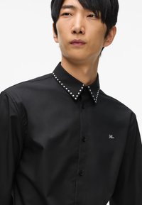 Fekete gombos ing, fitted fazonnal, szegecselt gallérral, sima textúrával és kis logóval a mellrészen.