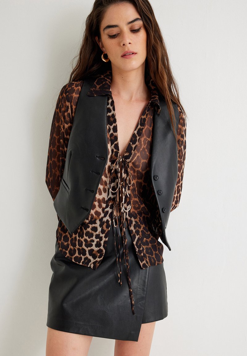 Czarna skórzana kamizelka nałożona na bluzkę w cętki w odcieniach brązu i czerni, z głębokim dekoltem i wiązaniem, w połączeniu z czarną skórzaną spódnicą mini.