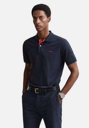 Marineblaues Polohemd mit roten Akzenten am Kragen, mit einem Logo auf der Brust. Kombiniert mit strukturierten marineblauen Hosen und einem schwarzen Gürtel.