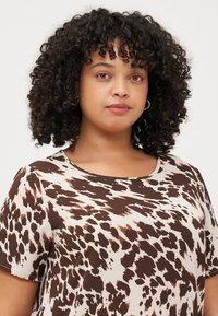 Young woman with curly hair models a short-sleeve shirt featuring a brown and cream animal print pattern. She has a neutral expression.
