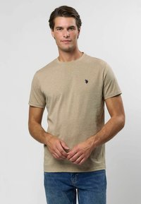 Khaki lühikeste varrukatega t-särk pehmest, tekstuuriga kangast, millel on ümmargune kaelus ja väike tumesinine logo vasakul rinnal. Kanditud siniste teksadega.