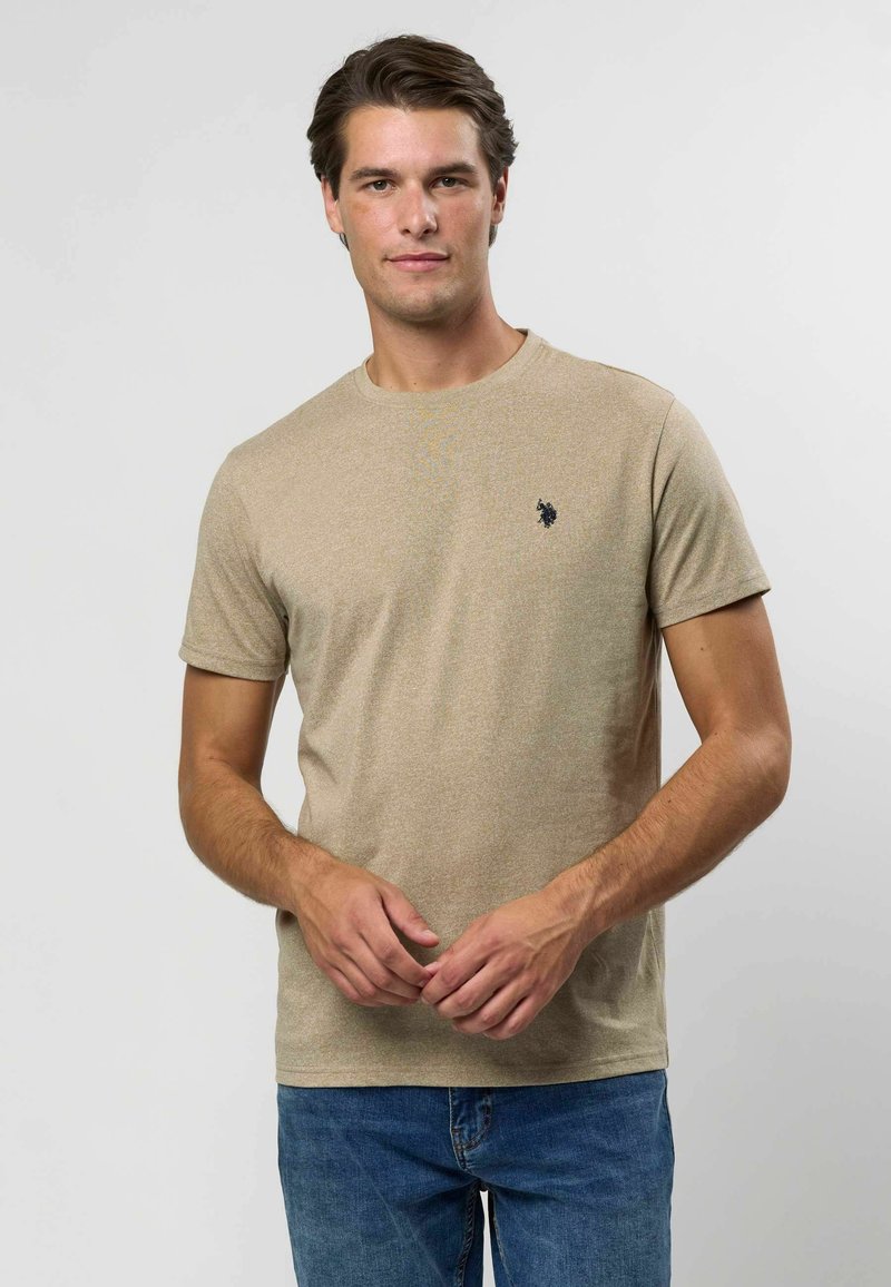 Khaki lühikeste varrukatega t-särk pehmest, tekstuuriga kangast, millel on ümmargune kaelus ja väike tumesinine logo vasakul rinnal. Kanditud siniste teksadega.