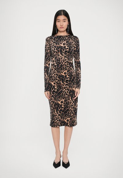 Een vrouw staat naar voren gekeerd in een jurk met lange mouwen en een leopardprint tot de knie, met zwarte puntige hakken, tegen een witte achtergrond.