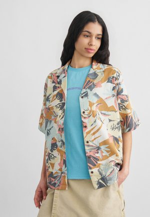 Jeune femme aux longs cheveux foncés portant une chemise bleue sous une blouse ample à manches courtes avec un motif floral, jupe beige, sur un fond uni.