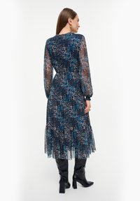 Lange, gelaagde jurk van doorzichtig materiaal met een blauw, zwart en wit bloemenpatroon. Lange mouwen en een getailleerde taille accentueren de silhouet.