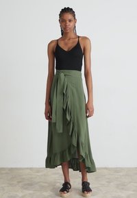 Top aderente nero abbinato a una gonna avvolgente verde oliva alta fino al ginocchio, con orlo arricciato e lacci in vita. Sandali neri.