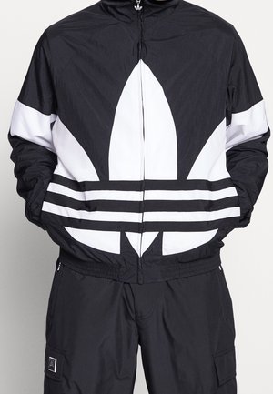 Personne portant une veste de sport Adidas noire et blanche avec un grand logo trèfle et un pantalon noir avec une poche latérale. Mains dans les poches de la veste.