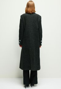 Cappotto lungo nero con una finitura testurizzata, dotato di una fessura sul retro e tre bottoni decorativi su ciascun polsino. Vestibilità su misura, si estende sotto le ginocchia.