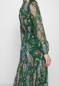 Vestido verde con mangas largas de gasa, cintura ceñida y un colorido patrón floral de paisley, llevado por una persona contra un fondo claro y liso.