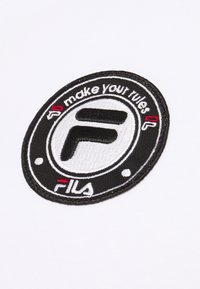 Logo ricamato in bianco e nero su uno sfondo di tessuto bianco, che presenta la lettera 'F' e il testo "fai le tue regole" e "FILA".