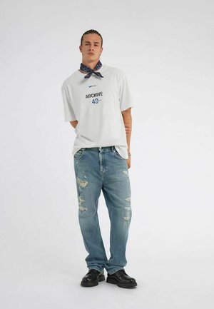 Joven de pie con las manos detrás de la espalda, vistiendo una camiseta blanca con gráfico, jeans azules desgastados, zapatos negros y pañuelo azul marino alrededor del cuello.