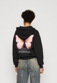 Osoba s kovrčavom kosom koja nosi crnu majicu s kapuljačom s velikim ružičasto-narančastim leptirskim motivom i tekstom na leđima, u kombinaciji s plavim trapericama.