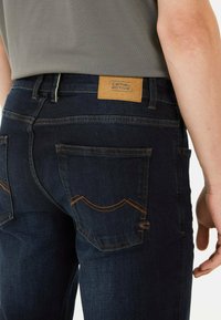 Donkerblauwe denim jeans hebben een leren patch met "camel active" branding, contrasterende oranje stiksels en gedetailleerde achterzakontwerpen.