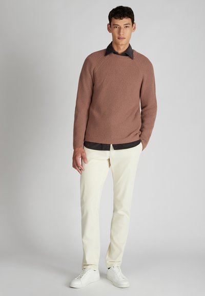 Man in een bruine geribde trui over een zwart shirt, crèmekleurige corduroy broek en witte sneakers, staand tegen een grijze achtergrond.
