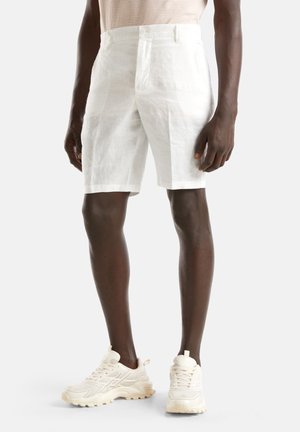 Homme portant un short blanc jusqu'aux genoux et des baskets couleur crème, debout devant un fond blanc.