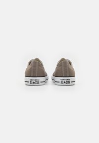 Baskets basses en toile marron avec des semelles en caoutchouc blanches, des rayures noires et un marquage "Converse All Star" au talon. Forme arrondie et détails cousus.