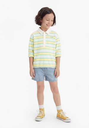 Smiling child with short curly hair wearing a pastel striped long-sleeve polo, denim shorts, white socks, and yellow high-top sneakers.
