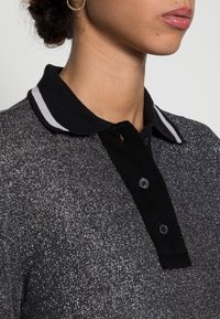 Femme portant un polo gris étincelant avec un col noir orné de rayures blanches, trois boutons noirs visibles et des boucles d'oreilles en or.