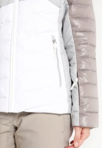 Veste matelassée blanche et grise avec poches zippées, portée sur un pantalon beige, montrant un gros plan du torse et du bras.