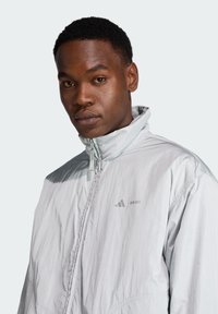 Chaqueta ligera de color gris claro, con un cuello alto, cierre frontal y un logo sutil en el pecho. Textura suave, diseño simple y ajuste moderno.