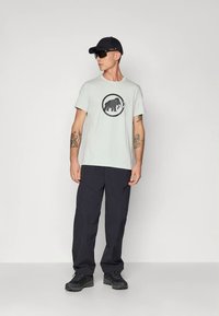 Vaaleanharmaa puuvillainen t-paita, jossa musta mammutti-grafiikka pyöreässä kuviossa. Yhdistetty tummiin cargo-housuihin ja mustiin lenkkareihin.