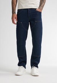 Uomo con braccia tatuate che indossa jeans blu scuro con toppe rinforzate sulle ginocchia e sneakers bianche, in piedi con una mano in tasca.