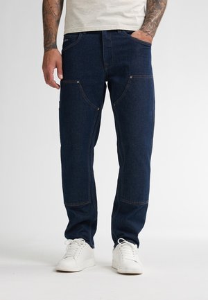 Man met getatoeëerde armen, gekleed in donkerblauwe jeans met versterkte kniestukken en witte sneakers, staand met één hand in zijn zak.