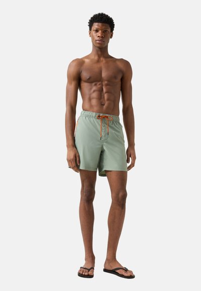 Homme torse nu portant un short de bain vert clair avec un cordon de serrage orange et des tongs noires, debout devant un fond uni.