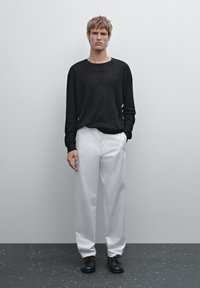 Sort ribbet strikket sweater, hvidt løstsiddende bukser, sorte sko med brune snørebånd. Model står foran en ensfarvet baggrund.