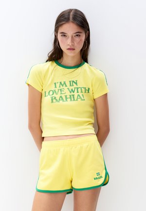 Geel kort mouw crop top met groene accenten en grafische tekst die zegt "IK BEN VERLIEFD OP BAHIA!" gecombineerd met bijpassende gele shorts met groene afwerking.