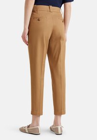 Kobieta w krótkich, beżowych spodniach z tylnymi kieszeniami na guziki, w beżowych płaskich, plecionych butach, stojąca na tle białym.