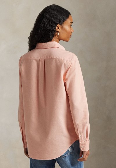 Femme aux cheveux bouclés et foncés, portant une chemise rose clair à manches longues et un jean bleu, vue de dos sur un fond neutre.