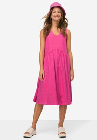 Abito fucsia senza maniche con scollatura a V, design a strati e silhouette fluida. Abbinato a un cappello rosa coordinato e sandali con zeppa beige.