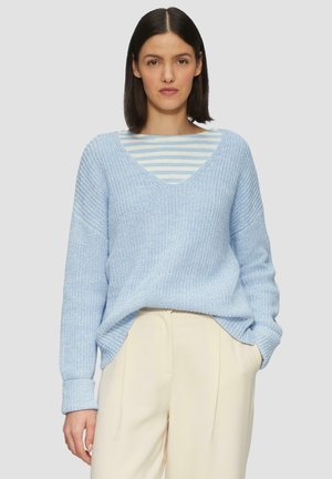 Hellblauer gestrickter Pullover mit V-Ausschnitt und gerippter Textur, über einem gestreiften Langarmshirt getragen. Kombiniert mit cremefarbenen Hosen.