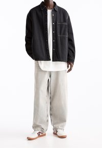 Sort denim skjortejakke med knaplukning og brystlomme, båret over en hvid skjorte. Lysegrå løse jeans og hvide sneakers med detaljer.