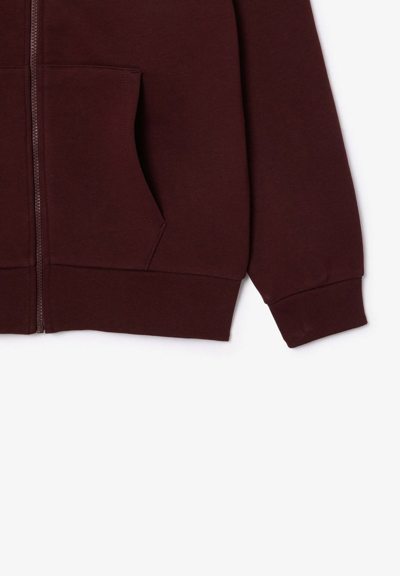 Sweat-shirt zippé bordeaux avec une poche avant, des poignets côtelés et une texture douce ; il dispose d'une fermeture éclair métallique et de détails de couture minimalistes.