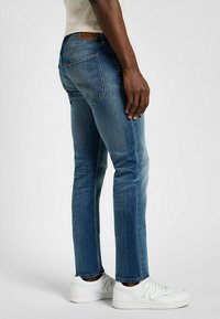 Slim-fit niebieskie dżinsy z delikatnym przetarciem, z tradycyjnymi tylnymi kieszeniami i brązowym skórzanym logo na pasku, w połączeniu z białymi trampkami.