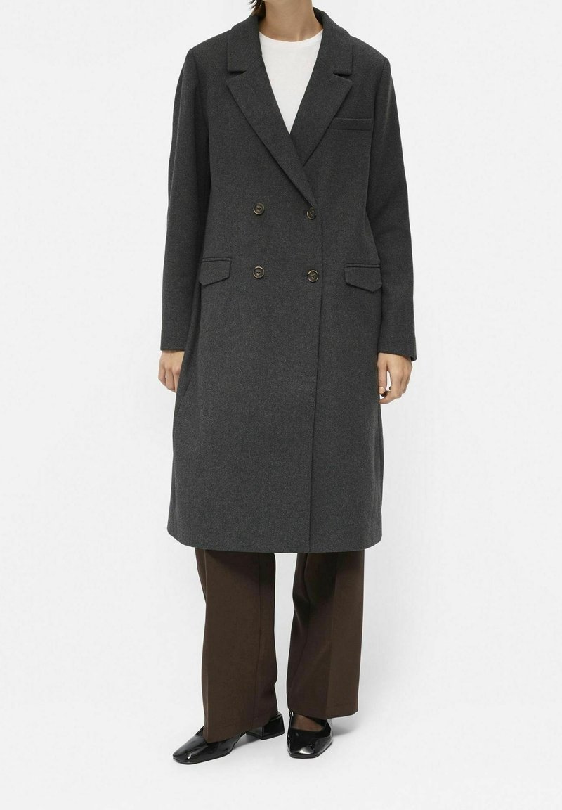 Personne portant un manteau gris foncé à double boutonnage sur un haut blanc, un pantalon marron à jambes larges et des chaussures noires à bout carré, debout sur un fond blanc.