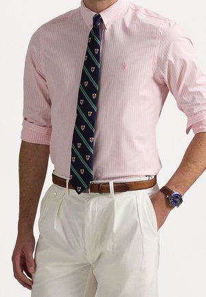 Homme en chemise rose à rayures avec manches retroussées, cravate bleu marine à rayures, pantalon blanc plissé, ceinture marron et montre bleue avec bracelet en cuir.