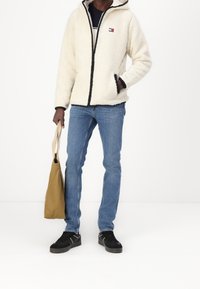 Vit fleecejacka med svart dragkedja och detaljer, blå jeans, svarta skor och en beige tote-väska. Jackan har en logotyp.