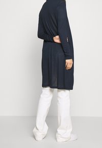 Robe longue à manches longues bleu marine avec une coupe décontractée, dotée d'un col rond et de fentes sur les côtés. Associée à un pantalon large blanc et des baskets blanches.