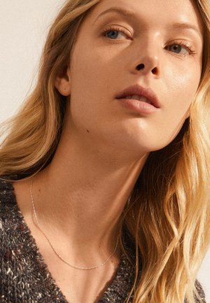 Sølvsnor halskæde med et delikat tvist design. Modellen ifører sig en struktureret, mørkt mønstret sweater med langt, løst hår.